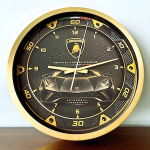 Könnte beinhalten: Eine runde Wanduhr mit goldfarbenem Rahmen und schwarzem Zifferblatt, das eine Lamborghini-Sportwagen-Grafik zeigt. Das Zifferblatt enthält das Lamborghini-Logo, den Text "DRIVEN BY A HIGHER STANDARD" und "AUTOMOBILI LAMBORGHINI" mit goldenen Ziffern.