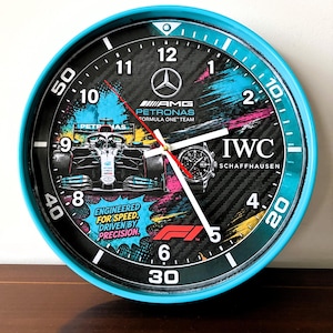 Op de afbeelding: Een ronde wandklok met een felcyaan frame en een zwarte wijzerplaat met koolstofvezelpatroon. Het toont een Mercedes-AMG Petronas Formule 1-auto, een IWC Schaffhausen horloge, het F1-logo en de tekst Engineered for Speed, Driven by Precision.