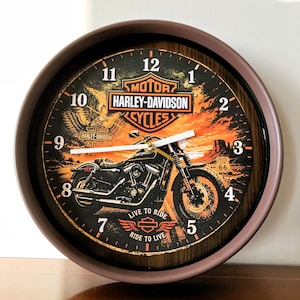 May include: A round wall clock with a brown frame featuring a Harley-Davidson motorcycle graphic, a sunset desert landscape, and a Route 66 sign. The clock face displays the text Harley-Davidson, Live to Ride, Ride to Live, and white hands.