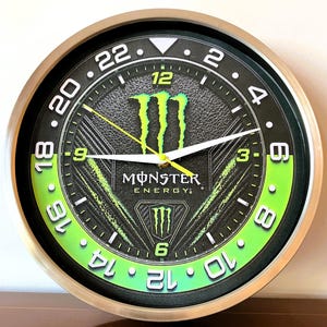 May include: A round wall clock with a metallic silver frame and a black textured face featuring the Monster Energy logo. The dial displays a 24-hour scale in white numerals, with a bright green accent section, white hour and minute hands, and a yellow second hand.