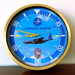 May include: A round wall clock with a gold-coloured frame and a bright blue face. The dial features the Lockheed Martin logo, a graphic of a C-130 Hercules aircraft, and the text C-130 HERCULES. It has yellow hour, minute, and second hands.