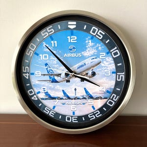 May include: A round wall clock with a metallic silver frame and a black outer ring featuring white minute markers. The clock face displays an Airbus A350 aircraft in flight against a blue sky with clouds, with additional aircraft on the ground below.