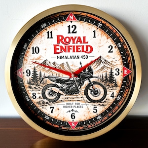 Op de afbeelding: Een ronde wandklok met een goudkleurige rand met een afbeelding van een Royal Enfield Himalayan 450 motorfiets, een berglandschap en kompasmarkeringen. De wijzerplaat toont de tekst Royal Enfield, Himalayan 450 en Built for Higher Places.