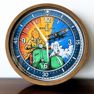 Peut inclure: Une horloge murale ronde avec un cadre en bois présentant un graphique coloré de Tintin sur un scooter Vespa vert avec Milou dans un side-car. Le cadran inclut le logo Breitling 1884, un avion et des marqueurs d'heure blancs sur un bord bleu.