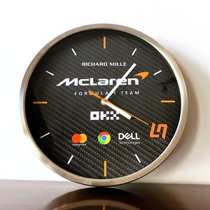 Könnte beinhalten: Eine runde Wanduhr mit silbernem Metallrahmen und einem Zifferblatt mit schwarzem Carbonfaser-Muster. Das Zifferblatt zeigt das McLaren Formula 1 Team Logo, Richard Mille, OKX, Mastercard, Chrome und Dell Technologies mit orangefarbenen und weißen Zeigern.