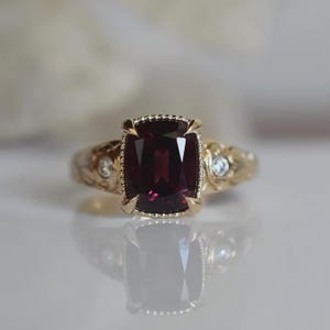 Puede incluir: Un anillo de oro con una gran piedra preciosa de color rojo intenso en talla cojín, engastada en un bisel con cuentas. La banda de oro amarillo presenta grabados florales intrincados y dos pequeños diamantes blancos redondos a los lados.