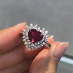 Puede incluir: Un anillo de tono plateado con una gran piedra preciosa de color rojo intenso en forma de corazón en el centro. La piedra central está rodeada por un doble halo de pequeñas piedras blancas redondas y claras en engaste de garras, con brillo.