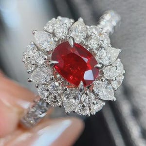 Puede incluir: Un anillo con una gran piedra preciosa de color rojo vibrante y corte ovalado en el centro, rodeada por un doble halo de numerosos diamantes blancos claros en forma de pera y corte redondo. La banda es de metal plateado pulido.