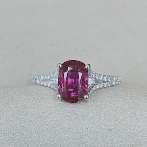 Puede incluir: Un anillo de tono plateado con una gran piedra preciosa de color rosa intenso y corte ovalado en el centro. La banda se divide en dos brazos delicados a cada lado, ambos incrustados con pequeñas piedras claras y brillantes a lo largo del aro.