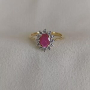 May include: A gold ring featuring a central oval-shaped pink gemstone surrounded by a halo of small, clear, round-cut stones. The band is thin and polished, providing a simple foundation for the prominent, multi-stone cluster setting.