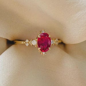 Puede incluir: Un anillo de oro con una gran piedra preciosa de color rojo intenso y corte ovalado como pieza central. El engaste está acentuado con pequeñas piedras blancas claras y redondas dispuestas en racimo a ambos lados de la piedra roja central.