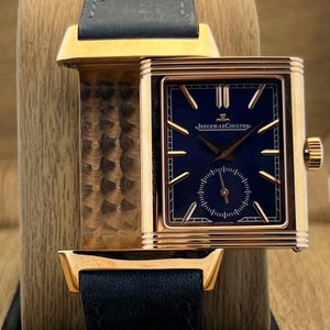 May include: A rectangular gold watch with a deep blue dial featuring the Jaeger-LeCoultre logo, gold hour markers, and a small seconds sub-dial. The watch has a dark blue leather strap and a metallic gold case with horizontal grooved edges.