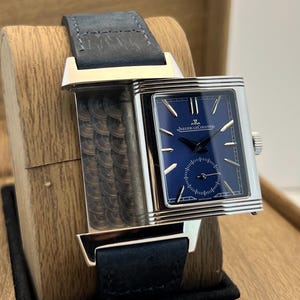 May include: A rectangular silver-tone wristwatch featuring a deep blue dial with silver indices and hands. The watch includes a small seconds sub-dial and the text Jaeger-LeCoultre. It is fitted with a dark blue textured leather strap.
