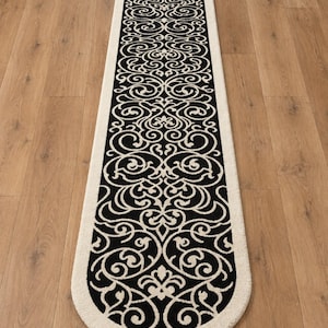 Peut inclure: Un long tapis de couloir étroit présentant un motif noir et crème. Le design se compose de volutes symétriques complexes et de motifs inspirés de fleurs. Le tapis possède des extrémités arrondies et une épaisse bordure crème entourant le centre noir.