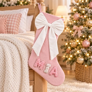 May include: A soft pink Christmas stocking featuring a large, white fabric bow at the top. The name MIA is appliqued on the front in padded letters, with the M and A in a pink and white gingham pattern and the I in a floral print fabric.