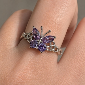 May include: A silver-toned ring featuring a butterfly design composed of four purple gemstones. The two upper wings are marquise-cut, while the two lower wings are round-cut. The band displays an intricate, openwork metal pattern on both sides.