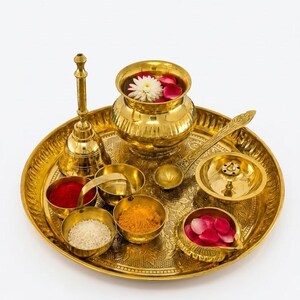 May include: A polished brass puja thali set featuring a large circular tray with intricate engravings, a central kalash pot filled with water and flower petals, a bell, a diya lamp, and several small bowls containing rice, turmeric, and red powder.