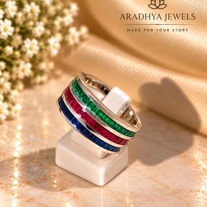 May include: A silver-toned band ring featuring three parallel rows of channel-set baguette-cut gemstones in green, red, and blue. The ring is displayed on a white stand with the text ARADHYA JEWELS MADE FOR YOUR STORY visible in the background.