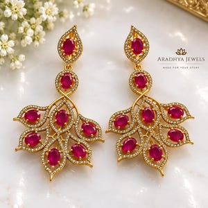 May include: A pair of gold-toned drop earrings featuring oval-cut vibrant pink stones arranged in a leaf-like pattern, accented with small white stones along the edges. The text Aradhya Jewels Made For Your Story is visible on the right side.
