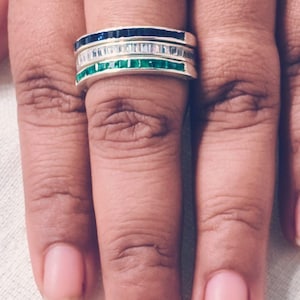 Puede incluir: Un conjunto de tres anillos metálicos en un dedo. El anillo superior tiene piedras azul oscuro en corte baguette, el anillo central contiene piedras blancas transparentes en corte baguette y el anillo inferior muestra una hilera de piedras verdes brillantes.
