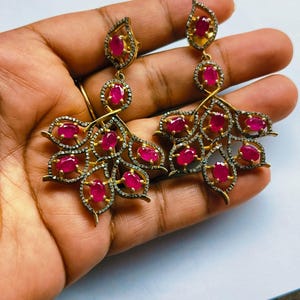 May include: A pair of gold-toned dangle earrings featuring oval-cut vibrant pink rubies set in a fan-like arrangement. Each earring is accented with small, dark-toned pavé stones along the edges of the metal frames, creating a detailed leaf-like pattern.