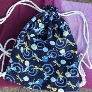 Puede incluir: Una mochila de cordón azul oscuro con un patrón repetitivo de libélulas coloridas, espirales azul claro y círculos verdes texturizados. La bolsa está confeccionada con una tela de superficie ligeramente cuadriculada y tiene cuerdas blancas.