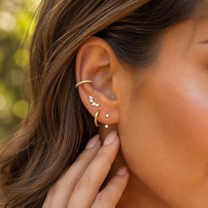 May include: A gold ear cuff with a textured surface, a gold stud earring featuring a cluster of marquise-cut clear stones, a small gold hoop earring, and a tiny gold stud earring with a single clear stone worn in a pierced ear.