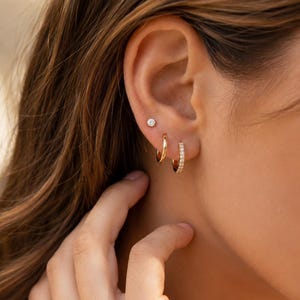 May include: A set of three gold-toned earrings consisting of one small stud with a clear stone, one plain polished gold hoop, and one gold hoop featuring a row of small clear stones along the front edge, all worn in a single ear lobe.