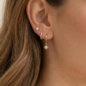 May include: A set of gold-toned earrings consisting of a small star-shaped stud with a central clear stone, a plain gold hoop, and a gold hoop featuring a dangling star-shaped charm with a central clear stone, all worn in a triple ear piercing.