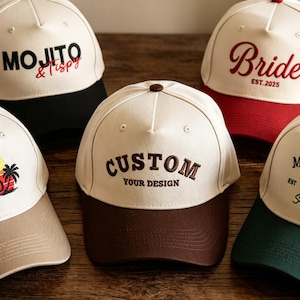 Peut inclure: Cinq casquettes de baseball avec des calottes blanc cassé et des visières colorées. Les casquettes présentent divers motifs brodés : 'MOJITO & Tipsy', 'Bride EST. 2025', 'SUN SALT SAND' avec un coucher de soleil, 'CUSTOM YOUR DESIGN' et 'MARGARITA EST 1998'.