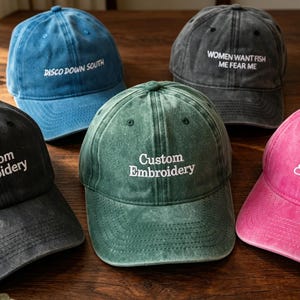 May include: Five baseball caps in blue, dark grey, green, black, and pink with a distressed finish. The caps feature white embroidered text including 'DISCO DOWN SOUTH', 'WOMEN WANT FISH ME FEAR ME', and 'Custom Embroidery' in various fonts.