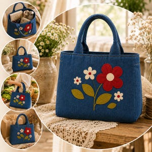 Op de afbeelding: Een blauwe denim draagtas met een grote rode bloemapplicatie met een wit hart, vergezeld door twee kleinere witte bloemen en groene bladeren. De tas heeft twee stevige handvatten aan de bovenkant en een gestructureerde rechthoekige vorm.