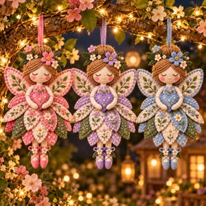 May include: Three hanging fabric fairy ornaments featuring brown yarn hair, floral crowns, and embroidered heart-shaped accessories. Each fairy has layered petal-shaped skirts in pink, purple, and blue, with matching ballet slippers and ribbon loops.