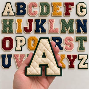 Pode incluir: Uma coleção de patches do alfabeto acolchoados em estilo universitário em várias cores, incluindo creme, azul marinho, rosa, verde floresta, amarelo mostarda e bordô. Cada letra apresenta um padrão de costura em diamante e uma borda lisa.