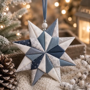 May include: A fabric star ornament featuring a geometric patchwork design in shades of navy blue, slate blue, and white. The star is constructed from triangular fabric segments with snowflake patterns and solid textures, hanging from a blue ribbon.