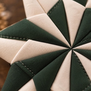 May include: A star-shaped fabric ornament featuring a geometric pattern of alternating dark forest green and cream-colored triangular fabric segments. The pieces are joined with visible stitching, creating a textured, quilted, multi-pointed star design.