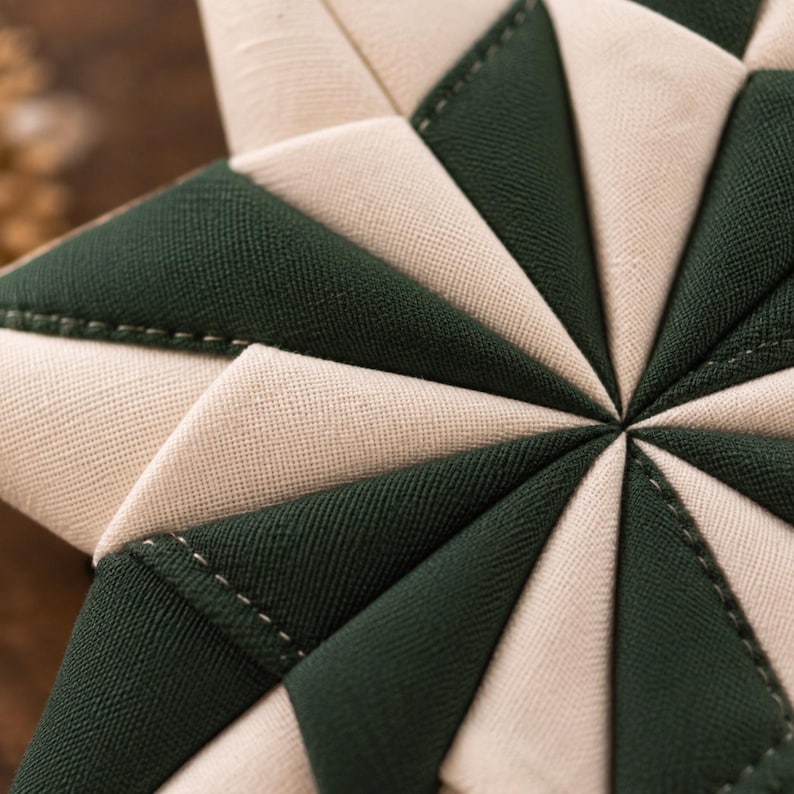 May include: A star-shaped fabric ornament featuring a geometric pattern of alternating dark forest green and cream-colored triangular fabric segments. The pieces are joined with visible stitching, creating a textured, quilted, multi-pointed star design.