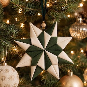 May include: A star-shaped fabric ornament featuring a geometric pattern of alternating dark green and cream-colored triangular panels. The ornament hangs from a dark green ribbon accented with a small wooden bead at the top point.