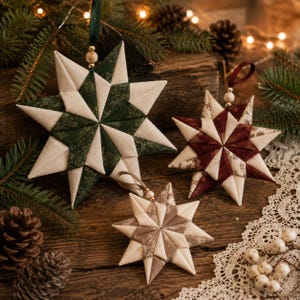 May include: Three fabric star ornaments featuring a geometric folded design. One large star is dark green and cream, a medium star is deep red and cream, and a small star is taupe and cream. Each star has a ribbon loop and a wooden bead at the top.