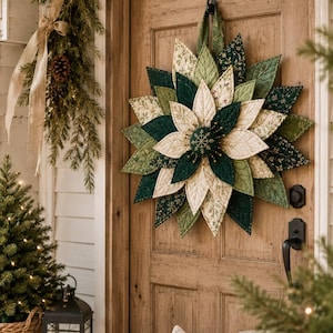 May include: A fabric wreath shaped like a large flower with layered, quilted petals in shades of cream, sage green, and dark forest green. The center features a dark green circular fabric piece with a gold embroidered snowflake design.