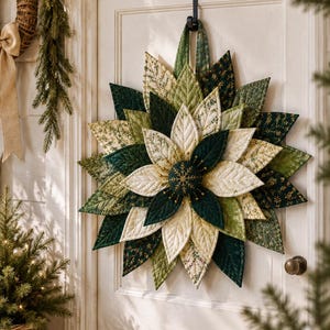 May include: A fabric star-shaped wall hanging featuring layered, quilted leaf-like petals in various shades of green, cream, and white. The center is a dark green circular piece with gold embroidered snowflake details and small gold bead accents.
