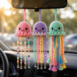 May include: Three hanging crochet jellyfish ornaments in pink, purple, and mint green. Each features a smiling face, beaded or crocheted tentacle strands, and clear teardrop-shaped charms at the bottom. The items are suspended by loops.