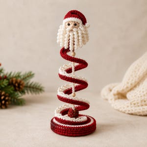 Crochet spiral Santa Christmas decoration displayed on a rustic wooden table with pine branches, gift box, and cozy cream knit sweater in the background
