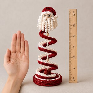 Puede incluir: Una figura de Pap&aacute; Noel tejida a ganchillo con gorro y barba roja y blanca, con un dise&ntilde;o de cuerpo en espiral que mide unos 25 cm de altura junto a una regla de madera. La base es circular, roja con borde blanco, y tiene ojos negros.