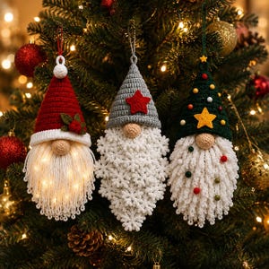 Pode incluir: Tr&ecirc;s ornamentos de gnomos de croch&ecirc; pendurados em uma &aacute;rvore. O gnomo da esquerda tem um gorro vermelho com azevinho e uma barba branca iluminada. O gnomo central tem um gorro cinza com uma estrela vermelha e uma barba de flocos de neve. O gnomo da direita tem um gorro verde com uma estrela amarela e pompons coloridos.