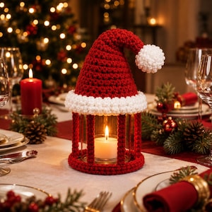 Könnte beinhalten: Ein zylindrischer Glaskerzenhalter, der mit einer rot-weißen Häkelhülle in Form einer Weihnachtsmannmütze ummantelt ist. Die Hülle hat eine rote Spitze mit einem weißen Bommel und einen weißen Rand. Eine brennende weiße Kerze steht im Glas.