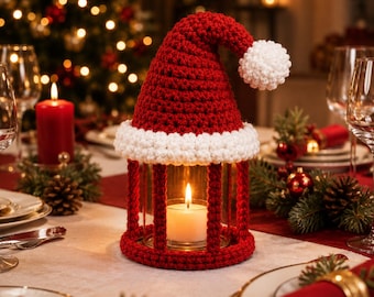 Pode incluir: Um suporte de vela cilíndrico de vidro envolto numa capa de croché vermelha e branca em forma de gorro de Pai Natal. A capa apresenta um topo vermelho pontiagudo com um pompom branco e uma aba branca. Uma vela branca acesa no interior.