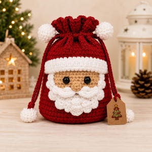 Peut inclure: Une pochette à cordon crochetée rouge et blanche représentant le Père Noël, avec une barbe, une moustache et un bord de bonnet blancs, des yeux en plastique noir et deux pompons blancs sur les cordons, avec une étiquette brune avec un sapin.