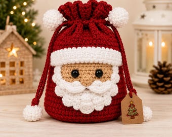 Pode incluir: Uma bolsa de cordão em crochê nas cores vermelho e branco com design de Pai Natal, apresentando barba, bigode e aba de gorro brancos, olhos de plástico preto e dois pompons brancos nos cordões, com uma etiqueta castanha com uma árvore.