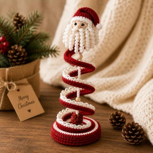 Puede incluir: Una figura de Pap&aacute; Noel tejida a ganchillo sobre una estructura en espiral roja y blanca. El Pap&aacute; Noel tiene un gorro rojo, barba blanca y ojos negros. La base en espiral est&aacute; hecha de hilo rojo y blanco con peque&ntilde;as cuentas de madera.